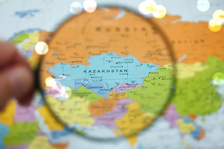 A hand holds a magnifying glass over Kazakhstan on a detailed world map. The map showcases vibrant colors representing various countries in Central Asia, emphasizing geography and exploration.